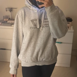 Grå hoodie från Emporio Armani EA7 - Snygg grå hoodie från Emporio Armani EA7 med stor logga framtill. Tröjan har huva med snörning, känguruficka och ribbade muddar. Perfekt för dig som gillar en clean och sportig stil. Mjuk och skön i bomullsblandning. Storlek XL men passar som en S. Är 158cm