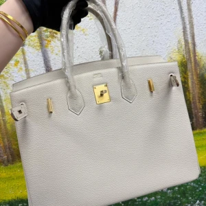 Beige handväska med gulddetaljer - Stilren beige handväska i grained skinn med klassisk fyrkantig form och två handtag. Väskan har guldfärgade metalldetaljer och lås framtill, samt en rymlig insida. Perfekt för dig som gillar en clean och lyxig look.