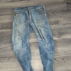 G-Star Raw ljusblå jeans - Säljer ett par ljusblå jeans från G-Star Raw med slitna detaljer och tvättad look. Jeansen har klassisk femficksdesign, raka ben och synliga sömmar längs benen för en cool streetstil. Perfekta för dig som gillar avslappnad och trendig vibe.