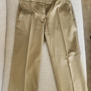 Beiga kostymbyxor från BRAX & more - Stilrena beiga kostymbyxor från BRAX & more med raka ben och snygga pressveck. Byxorna har två fickor fram och två bakfickor med lock. Tillverkade i ett mjukt bomullsmaterial som känns skönt mot huden. Perfekta för en clean och klassisk look. Ser ljusare ut på bilden men är khaki/ oliv grön.