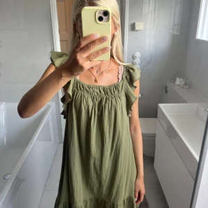 Grön volangklänning med ärmlös design - Supersöt ljusgrön klänning med volangdetaljer vid axlarna och lös passform. Ärmlös modell med rynkad överdel och flowy kjol, perfekt för varma dagar. Tillverkad i mjukt bomullstyg som känns skönt mot huden.