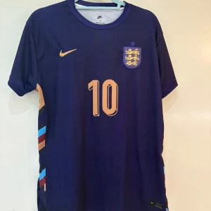 Bellingham England Euro2024 - Säljer en snygg Jude Bellingham (nummer 10)  England bortatröja från Euro 2024. Tröjan är i den stilrena mörklila designen med guldfärgade detaljer. Strlk M, oanvänd och riktigt härlig sommartröja!
