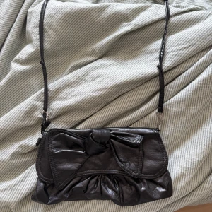 Svart axelväska med rosett - Säljer en svart axelväska i skinnimitation med snygg rosettdetalj framtill. Väskan har rynkade detaljer nedtill och ett avtagbart axelband. Stängs med magnetknapp och har ett foder i svart tyg. Perfekt storlek för det viktigaste.