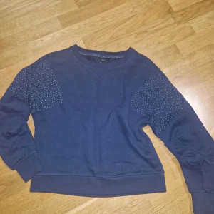 Mörkblå sweatshirt med pärldetaljer ONLY - Mörkblå sweatshirt från ONLY i storlek XS. Tröjan har långa ärmar och coola pärldetaljer på axlarna som ger en extra touch. Mjuk och skön bomullskänsla, perfekt till jeans eller kjol. Rund halsringning och ribbade muddar vid ärmslut och nederkant.