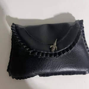 Svart plånbok i skinn med detaljer - Cool svart plånbok i skinn med dekorativ kant och unik metallknapp framtill. Plånboken har flera fack och dragkedjor på insidan för mynt och kort. Perfekt storlek för att ha med sig överallt och ger en edgy vibe till din stil.