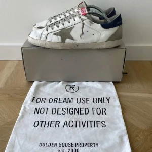 Golden Goose Superstar sneakers vit/grå - Golden Goose Superstar sneakers i vitt skinn med grå mockadetaljer och blå häl. Klassisk stjärna på sidan, snörning och platt sula. Snyggt slitna för en cool streetstyle-look. Perfekta för dig som gillar exklusiva och trendiga sneakers.