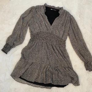 Svartvit kortklänning från Zara - Supersöt kortklänning från Zara i svartvitt småmönstrat tyg. Klänningen har v-ringning, långa ärmar med resår vid handlederna och smock i midjan för en snygg passform. Volang nertill och lätt, luftig.