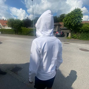 Vit vindjacka från Moncler med huva - Snygg vit vindjacka från Moncler med dragkedja framtill, justerbar huva och svarta detaljer vid ärmslut och fickor. Jackan har Moncler-logga på ärmen och en clean, sportig look som passar perfekt till streetwear.
