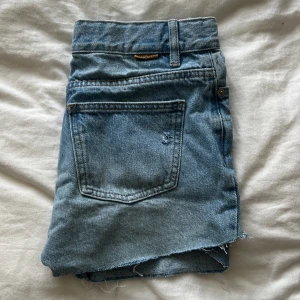 Ljusblå jeansshorts  - Säljer ett par ljusblå jeansshorts från Studio Total med råa, fransiga kanter och slitna detaljer. Klassisk femficksmodell med dragkedja och knapp framtill. Perfekta för en avslappnad och cool stil under varma dagar.