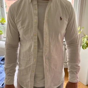 Vit slim fit skjorta från Ralph Lauren - Klassisk vit skjorta från Ralph Lauren i slim fit-modell. Knappar framtill samt det ikoniska broderade Polo-loggan på bröstet. Perfekt för dig som gillar en stilren och tidlös look.