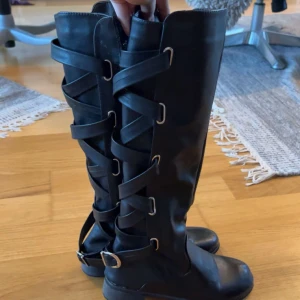 Svarta höga stövlar med snörning - Snygga svarta stövlar i skinnimitation med dekorativ snörning och spännen längs hela skaftet. Platt sula och rund tå ger en cool och bekväm look. Perfekta för att lyfta din outfit med en edgy touch.