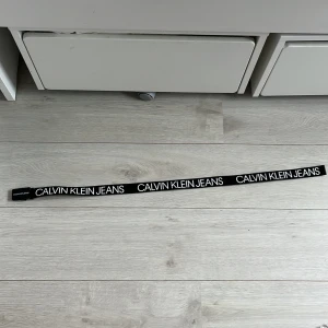 Svart Calvin Klein Jeans bälte L/XL - Snyggt svart bälte från Calvin Klein Jeans med tydlig vit logga tryckt längs hela bandet. Bältet har en modern, rak form och svart spänne. Perfekt accessoar för att lyfta din outfit med en trendig streetstyle-vibe.