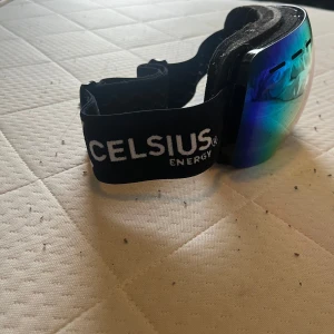 Celsius Energy skidglasögon  - Säljer ett par feta skidglasögon från Celsius Energy med svart justerbart band och vit logga. Linsen är spegelblå med inslag av gult och grönt, vilket ger en riktigt cool look i backen. Skumkant för extra komfort och bred passform. Perfekta för vinterns äventyr! Pris ej hugget i sten vid snabb affär