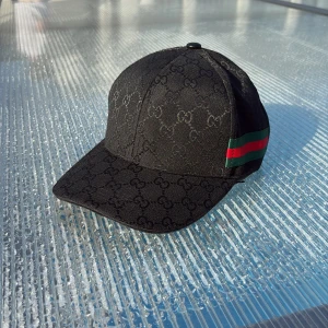 Svart Gucci keps med GG-mönster - Exklusiv svart keps från Gucci med ikoniskt GG-monogram över hela kepsen. Snygg detalj med grönt och rött band på sidan. Justerbar rem baktill. Tillverkad i 90% merinoull och 10% kashmir för lyxig känsla.