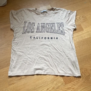 Beige t-shirt Los Angeles FB Sister - Beige t-shirt från FB Sister med stor svart text 'LOS ANGELES CALIFORNIA' på framsidan. Klassisk rund hals och korta ärmar. Skön och avslappnad passform, perfekt till jeans eller shorts. Tillverkad i mjuk bomull.