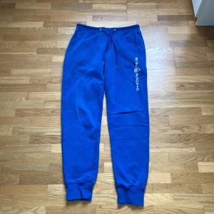 Blå mjukisbyxor Sail Racing S - Blå mjukisbyxor från Sail Racing med vit logga längs benet. Byxorna har snörning i midjan, muddar vid bensluten och fickor fram samt en bakficka. Sköna och sportiga, perfekta för chill eller träning.