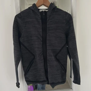 Svart hoodiejacka från Sail Racing S - Svart hoodiejacka från Sail Racing i sportig stil. Jackan har dragkedja framtill, huva och coola meshdetaljer på sidorna. Materialet är syntet och jackan har en diskret mönstrad struktur. Korsad i ryggen.