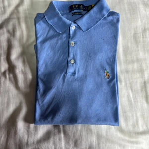 Blå pikétröja från Ralph Lauren - Snygg ljusblå pikétröja från Ralph Lauren med klassisk krage och tre knappar framtill. Tröjan har det ikoniska broderade polospelar-loggan på bröstet och är gjord i mjuk bomull. Perfekt för en clean och stilren look.