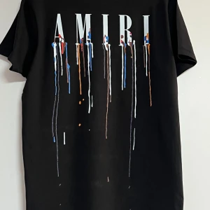 Svart AMIRI t-shirt med färgstänk - Svart t-shirt från AMIRI med stort tryck på ryggen och mindre tryck fram. Trycket består av AMIRI-loggan och färgstänk i vitt, blått, rött och orange som rinner ner. Klassisk passform och rund hals, tillverkad i mjuk bomull.