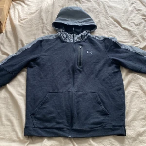 Under Armour huvtröja - Snygg grå hoodiejacka från Under Armour med grå detaljer på huva och axlar. Jackan har dragkedja hela vägen, en bröstficka med dragkedja och två stora fickor framtill. Perfekt för dig som gillar sportig stil och vill ha något bekvämt och funktionellt.