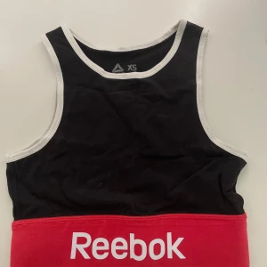 Sporttopp - Sporttopp från Reebok i svart och rött med vit kant runt. Toppen har en Reebok-logga framtill och är tillverkad i mjuk bomull, perfekt för träning eller chill dagar.