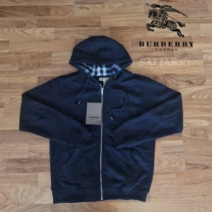 Svart Burberry Hoodie - Letar du efter en stilren och lyxig hoodie? Denna helt nya grå Burberry-hoodie kombinerar elegans med hög komfort. Den har Burberrys ikoniska rutmönster i fodret och en diskret logga på bröstet för en exklusiv touch. Tillverkad av mjukt, slitstarkt material och utrustad med dragkedja samt huva – både praktisk och stilfull.  📌 Skick: Ny med taggar. 📦 Snabb leverans – skickas samma dag! 