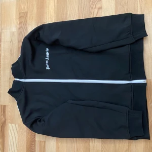 Palm Angels svart track jacket - Svart track jacket från Palm Angels med vit dragkedja och vita ränder längs ärmarna. Jackan har hög krage och broderad Palm Angels-logga på bröstet. Tillverkad i mjukt syntetmaterial, perfekt för en sportig och trendig look.