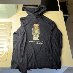 Mörkblå tshirt/hoodie från Ralph Lauren - Säljer en mörkblå tshirt/hoodie från Ralph Lauren med en cool Polo Bear-design på framsidan. Tröjan har långa ärmar och en justerbar huva med dragsko. Perfekt för en avslappnad stil!
