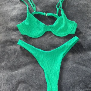 Grön bikini med ribbad textur - Snygg grön bikini med ribbad textur och justerbara axelband. Toppen har en dekorativ spänne framtill och bygel för extra stöd. Perfekt för en dag på stranden eller vid poolen.