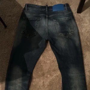 Mörkblå jeans med slitningar - Snygga mörkblå jeans med coola slitningar och färgstänk. De har en klassisk femficksdesign och knappgylf. Perfekta för en avslappnad stil.