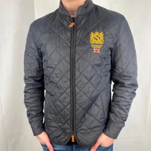 Svart quiltad jacka från Morris - Snygg svart quiltad jacka från Morris med dragkedja och broderad logotyp på bröstet. Jackan har en stilren design med långärmade ärmar och en liten ståkrage. Perfekt för en stilren look.