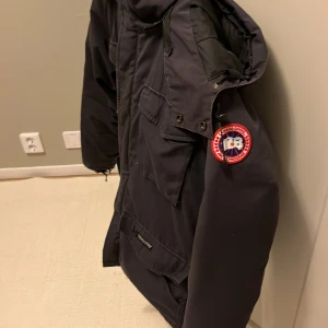  Canada Goose jacka - Säljer en svart vinterjacka från Canada Goose med lång ärm och flera praktiska fickor. Jackan har en hög krage och en tydlig logotyp på ärmen. Perfekt för kalla dagar. Hör av er vid minsta frågan!