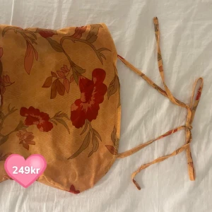Blommig tubtopp i orange och rött - Säljer en snygg blommig tubtopp i orange med röda blommor. Toppen har smala axelband som knyts i nacken och en croppad passform. Perfekt för sommardagar! Storlek S men passar XS!