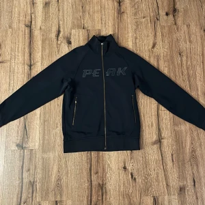 Mörkblå jacka från Peak Performance - Snygg mörkblå jacka från Peak Performance med dragkedja framtill och två fickor med dragkedjor. Jackan har en stilren design med märkesnamnet diskret tryckt på bröstet. Perfekt för en sportig look!