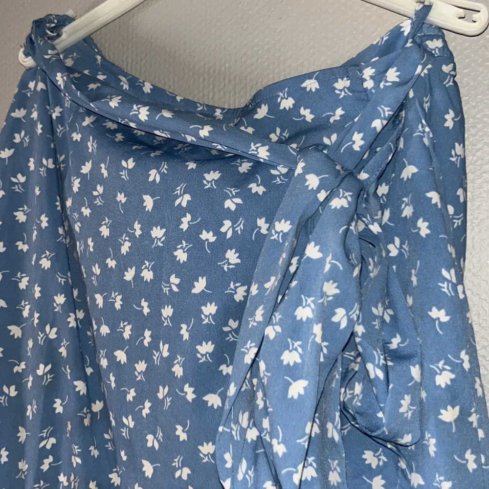Säljer blå kjol med vita blommor från ONLY. Kjolen har en omlott-design med volangdetaljer och ett knytband i midjan!. Hameet.
