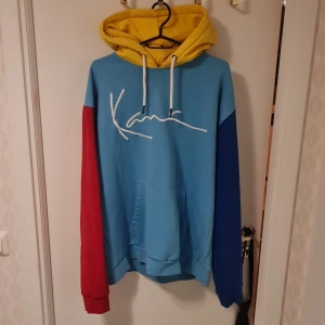Flerfärgad hoodie från Karl Kani - Säljer en cool flerfärgad hoodie från Karl Kani. Den har en gul huva, blå kropp och ärmar i rött och blått. Stort vitt tryck på framsidan och praktisk magficka. Perfekt för en färgglad stil!