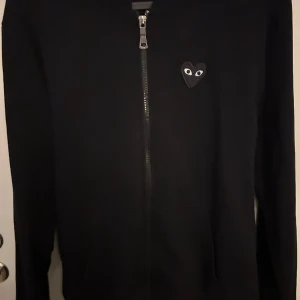 Svart hoodie från Comme des Garçons - Säljer en svart hoodie från Comme des Garçons med dragkedja och ett ikoniskt hjärtmärke med ögon på bröstet. Baksidan har ett större hjärtmärke. Perfekt för en stilren look. Storlek S men passar även bra på XS