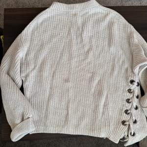 Beige stickad tröja med snörning - Säljer en beige stickad tröja med unik snörning på sidan. Tröjan har långa ärmar och en ribbad struktur som ger en stilren look. Perfekt för kyligare dagar.