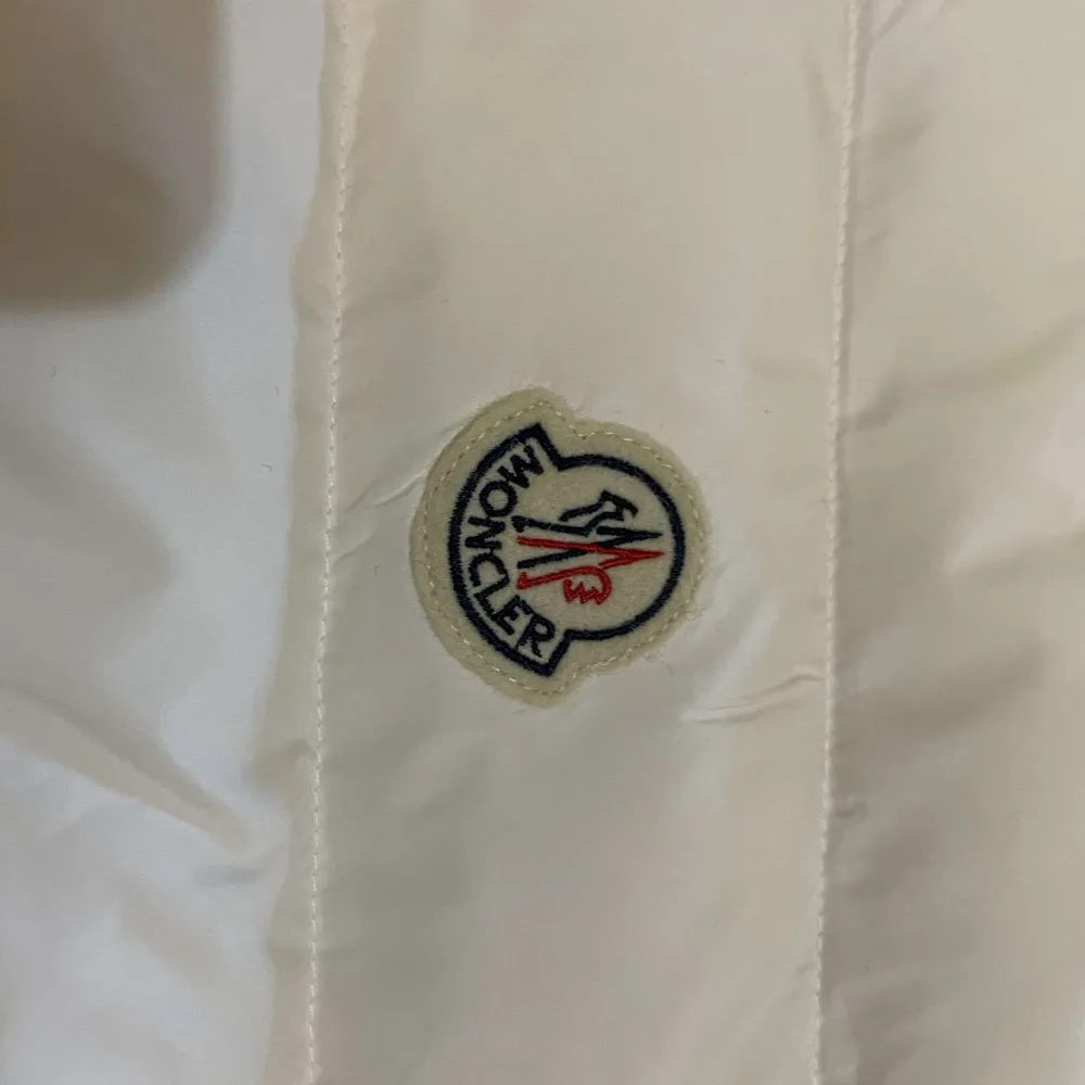 Snygg vit dunjacka från Moncler med dragkedja och knappar. Jackan har ett stilrent märke på bröstet och ribbade muddar. Perfekt för kyliga dagar när du vill hålla stilen. 🏔️. Takit.