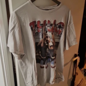 Vit T-shirt med tryck från Bershka - Säljer en vit T-shirt från Bershka med ett stort tryck på framsidan som säger 'Thug Life' och en bild. Perfekt för dig som gillar streetwear och vill ha en avslappnad stil. T-shirten är kortärmad och gjord i bomull.