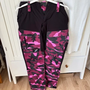 Rosa och svarta camo byxor från RevolutionRace - Coola byxor från RevolutionRace i rosa och svart camouflagemönster. Byxorna har flera praktiska fickor och en bekväm passform. 
