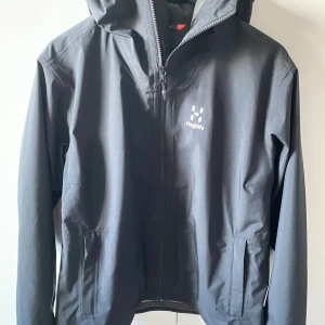 Svart jacka från Haglöfs - Säljer en svart jacka från Haglöfs med dragkedja och huva. Jackan har en stilren design med två fickor framtill och är perfekt för utomhusaktiviteter.🩷