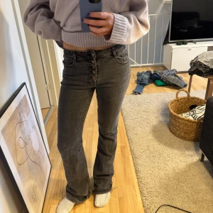 Grå bootcut jeans med knappar - Snygga grå bootcut jeans med hög midja och knappar framtill. Perfekta för en avslappnad stil med en twist. De har en klassisk femficksdesign och en bekväm passform.