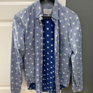 Blå stjärnmönstrad skjorta från Our Legacy - Snygg blå skjorta från Our Legacy med ett coolt stjärnmönster. Skjortan har långa ärmar och knappar framtill. Perfekt för att sticka ut med en unik stil.