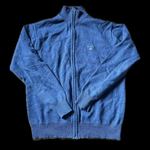 Blå kofta från Gant - Säljer en stilren blå kofta från Gant med dragkedja framtill. Tröjan har långa ärmar och ribbade muddar vid ärmslut och nederkant. Perfekt för kyliga dagar!