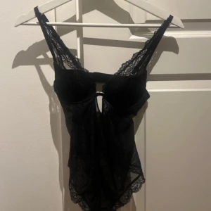 Svart spetsbody - Snygg svart spetsbody med smala axelband och dekorativ spetsdetalj. Den har en djup urringning och en elegant design som framhäver figuren. Perfekt för en stilfull look.