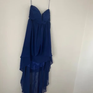 Blå axelbandslös balklänning - Säljer en elegant blå axelbandslös klänning med v-ringad design och asymmetrisk längd. Perfekt för festliga tillfällen. Klänningen har en luftig och flödande stil som ger en vacker siluett.