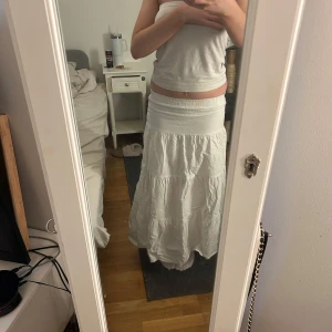 Vit långkjol - Säljer en vit långkjol med elastisk midja. Kjolen har en luftig och bekväm passform, perfekt för varma dagar. Den är tillverkad i ett lätt material som ger en skön känsla.