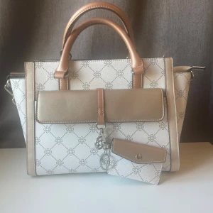 Vit och beige handväska med mönster - Elegant handväska i vitt och beige med ett subtilt mönster. Väskan har både handtag och en avtagbar axelrem för flexibilitet. Framsidan pryds av en dekorativ detalj och en matchande liten plånbok. Insidan är rymlig med dragkedja för säker förvaring.
