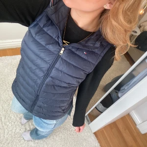 Mörkblå väst från Tommy Hilfiger - Snygg mörkblå dunväst från Tommy Hilfiger med dragkedja och huva. Perfekt för kyligare dagar och ger en stilren look. Västen har en liten logga på bröstet och är lätt att matcha med olika outfits. Nypris: 1800kr